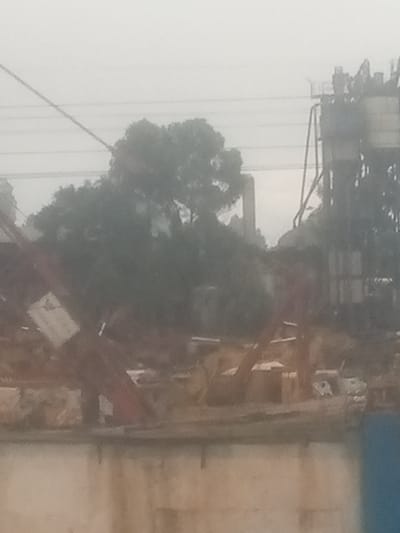 This image, captured in Sigona ward, Kenya, presents a hazy, industrial outdoor scene under an overcast sky. In the upper right background, a tall industrial structure, possibly a concrete plant or factory, is visible, characterized by multiple levels, vertical supports, and cylindrical silos or tanks. Power lines traverse horizontally across the upper portion of the frame, passing above a dense cluster of dark green trees occupying the mid-ground. In the lower mid-ground and foreground, fragmented reddish-brown and dark-colored industrial equipment, potentially parts of a crane or construction machinery, are discernible. One piece of equipment on the left features a rectangular white element, possibly a sign or panel. The immediate foreground consists of a light-colored, stained wall, likely concrete, extending across the bottom width of the image. A narrow, dark blue vertical object is partially visible on the far right edge of the foreground. No individuals are visible, and no specific actions or interactions are depicted. The overall environment suggests an active or recently active industrial site with some natural vegetation.