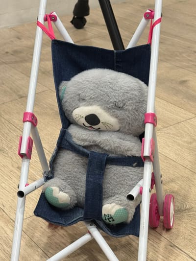 A close-up, high-angle shot shows a gray teddy bear with its eyes closed, sleeping in a miniature blue denim stroller. The stroller has a white metal frame and pink accents on its wheels and attachment points. The teddy bear is nestled in the denim seat, with its arms crossed. The background is a light-colored wooden floor. The image is well-lit and in focus. There is no information about the location of Krasnyi Krym, Russia in the image.
