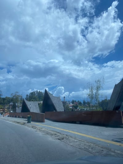The image captures a view from a street in Parapat, Indonesia, looking towards a waterfront area with distinctive architecture. Several triangular-roofed structures, likely part of a resort or public facility, dominate the mid-ground. These structures feature dark-colored, angular roofs with visible wooden beams and appear to be constructed with contemporary design elements.
In front of these structures is a wide, paved walkway or promenade, bordered by a low, dark brown barrier. A single green trash can is visible on the walkway near the left side of the image. The walkway is marked with tactile paving for the visually impaired, indicated by rows of raised dots, and a yellow line running parallel to it.
The background reveals a hillside densely covered with trees and a scattering of buildings, suggesting a town or residential area nestled into the landscape. The sky above is a dramatic mix of bright blue patches and heavy, grey clouds, indicating a dynamic weather system. A thin, bare tree with drooping branches is positioned between two of the prominent structures, its delicate form contrasting with the solid architecture.
The foreground consists of a grey asphalt road, with the edge of a vehicle visible at the bottom of the frame, suggesting the image was taken from a car. The road surface shows some signs of wear and tear, with cracks and debris along the edge. The overall impression is of a scenic, possibly tourist-oriented location within Parapat, characterized by its unique architectural style and natural surroundings under a cloudy sky.