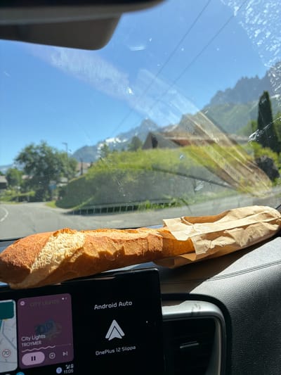 A close-up, forward-facing, eye-level shot captures a baguette resting on a car's dashboard. The bread, a light brown crust with a white interior, is partially wrapped in a brown paper bag and extends across the frame from left to right. Below the baguette, a car's infotainment screen displays "Android Auto" and music information for "City Lights" by Troymar. The background, seen through the windshield, reveals a bright blue sky with some clouds, lush green trees, houses, and snow-capped mountains, consistent with the location of Les Houches, France. The scene is illuminated by natural daylight.