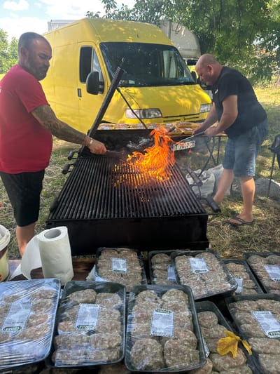 Two men are in an outdoor setting, preparing to grill a large quantity of raw meat patties. One man, wearing a black t-shirt and denim shorts, is using tongs to place patties on a large grill. Flames are visible beneath the grill grates. The second man, in a red t-shirt and black shorts, is standing near the grill, holding a tool that appears to be a grill brush.

In the background, a bright yellow van is parked. To the right of the grill, several plastic trays of raw meat patties are stacked, each wrapped in plastic and labeled with Cyrillic text. A roll of paper towels is also visible next to the grill.

The scene suggests a preparation for a barbecue or a similar outdoor food event. The lighting and shadows indicate it is daytime, likely sunny. The environment appears to be grassy with some trees in the background, suggesting a rural or park-like location. The text on the meat labels is in Cyrillic, consistent with the provided location context of Lovets, Bulgaria.