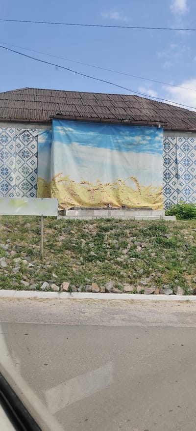 The image shows a building with a corrugated roof and a wall decorated with a blue and white patterned tile mosaic on the left side. A large banner depicting a landscape of wheat fields under a blue sky with clouds is hung across the central part of the wall. In front of the building, there is a sloped area covered with grass and rocks, with a white rectangular sign on a pole to the left. The foreground shows a paved road. The scene is located in Kolomak, Ukraine.