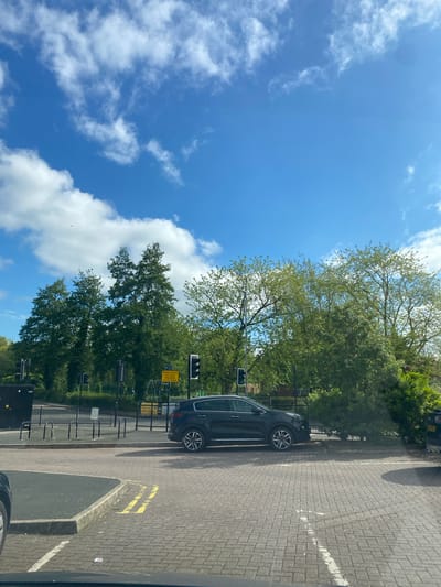 The image shows an outdoor scene in Telford, United Kingdom, on a sunny day with a blue sky dotted with scattered white clouds. A dark-colored SUV is parked on a brick-paved area next to a sidewalk. Adjacent to the sidewalk, there's a metal fence, likely delineating a park or green space. The background features a row of trees with green foliage. Traffic lights and a yellow traffic sign are visible behind the car. Double yellow lines are painted on the brick pavement next to the curb.