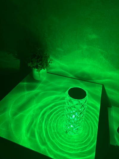 In Dmitriyevskaya, Russia, an indoor scene is depicted under strong green illumination. On a flat, light-colored surface, presumably a table, a faceted, cylindrical crystal lamp is positioned on the right, emitting a bright green light. This light casts distinct concentric ripple patterns onto the table surface and wavy, abstract patterns onto the textured wall in the background. To the left on the same surface, a small potted plant with dark green foliage rests within a light-colored pot, its shadow extending upwards on the wall. No individuals are present in the scene.