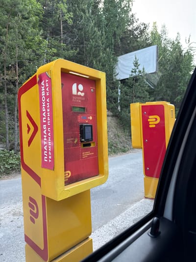 The image shows a payment terminal for parking, likely located at the entrance or exit of a parking area. The machine is predominantly yellow with red accents and features a prominent logo. The terminal accepts card payments as indicated by the inscription "ПЛАТНАЯ ПАРКОВКА ТЕРМИНАЛ ПРИЕМА КАРТ" (Paid Parking Card Acceptance Terminal). There is also a card reader and slots for inserting parking cards. Additional text in Russian is visible, including "ВСТАВЬТЕ ПАРКОВОЧНУЮ КАРТУ" (Insert Parking Card), "ПОДНЕСИТЕ КАРТУ ДОСТУПА" (Present Access Card), "СПРОСИТЬ" (Ask), and "Золотой пляж" (Golden Beach). The terminal is located near trees and foliage, with a white structure visible in the background through the foliage. The scene is viewed from inside a car. Dom Otdykha Zolotoy Plyazh, Russia is the location in the city.