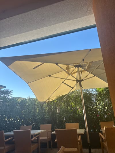 This media file depicts an outdoor seating area at a cafe or restaurant. The primary subjects are tables and chairs, most of which are made of wicker or a similar woven material. An open, light-colored patio umbrella is visible, extending upwards and casting shadows. The scene is set against a backdrop of lush green trees and a clear blue sky, suggesting it is daytime. The lighting indicates a sunny day, likely in the late morning or afternoon.
The setting appears to be an outdoor patio or garden area, possibly in a European location given the mention of Gebersdorf, Austria. There are no people visible in the image, suggesting the establishment is either closed or the patrons are not currently in the frame. The focus is on the empty seating arrangements, creating a sense of quiet anticipation or a peaceful moment.
Notable details include the texture of the chairs and tables, the structure of the umbrella's framework, and the dense foliage of the trees. The angle of the shot, looking upwards towards the umbrella and sky, provides a unique perspective on the outdoor furniture and the natural surroundings. The overall impression is one of a pleasant, sunlit day in a relaxed outdoor environment. There is no visible text in the image.