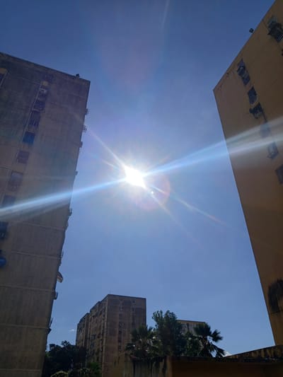 The image depicts an outdoor urban scene in Puerto Ordaz, Venezuela, characterized by a clear, bright blue sky and harsh sunlight. Two tall, weathered apartment buildings dominate the foreground, angled towards the viewer from the left and right sides of the frame, respectively. The building on the left appears to be concrete-gray, with numerous visible air conditioning units and balconies. The building on the right is a pale yellow, also showing signs of age, with similar architectural features. The sun is positioned prominently in the upper center of the image, causing lens flare and bright streaks of light that extend across the sky and the buildings. In the distance, a third, slightly shorter apartment building can be seen, along with lush green trees and palm fronds, suggesting a tropical or subtropical environment. The overall impression is one of a sunny day in a densely populated urban area.