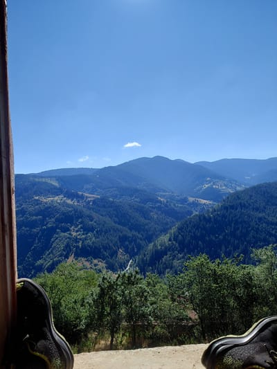 An upward angle shot shows a pair of black and yellow sneakers resting on a dirt ground, framed by a wooden structure on the left. Beyond the sneakers, a vast expanse of rolling, forest-covered mountains stretches across the horizon under a clear blue sky with a single small cloud. The scene appears to be a scenic overlook in Dryanovets, Bulgaria, offering a panoramic view of the natural landscape.