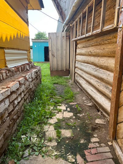 The image depicts a narrow pathway between two rustic wooden structures and a brick wall, likely in a rural or semi-rural setting within Большой Кукмор, Russia. The pathway is uneven, paved with broken pieces of brick and stone, and overgrown with green vegetation, suggesting it is not heavily trafficked. To the left, a yellow-painted wooden building with white siding at its base rises prominently. To the right, a log cabin-style structure with a visible wooden ladder or shelf system along its upper exterior wall forms the boundary of the pathway. A wooden door, partially ajar, is visible on the log cabin. In the background, a small blue shed is visible, adding to the sense of a utilitarian or residential yard. The overall impression is one of a simple, weathered, and somewhat unkempt outdoor space. There are no people or animals present in the image. The lighting suggests it is daytime, with sunlight casting shadows.