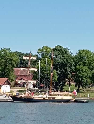 This media file depicts a picturesque waterside scene in Sztynort, Poland. The central subject is a large sailboat docked along a grassy shoreline, its masts reaching towards a clear blue sky. The boat has a classic wooden hull with a cream-colored deck and dark blue accents. Several people are visible on the deck, suggesting it is a lively day for sailing or tourism. A prominent red and white flag is flying from the boat's mast.
Behind the boat, lush green trees create a dense backdrop, interspersed with glimpses of buildings, including a red-roofed structure. The water in the foreground is a calm, muted blue, reflecting the bright sky. The scene appears to be set on a sunny day, likely in the late morning or early afternoon given the quality of the light. There are no visible inscriptions or text in the image. The overall atmosphere is one of peaceful recreation and natural beauty, characteristic of a summer day at a lakeside or harbor.