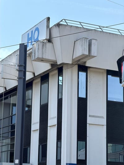The image shows a modern building with a distinctive black and white facade. The building has a series of horizontal black ribbed panels interspersed with white sections. Numerous rectangular windows are visible, some of which reflect the sky. The building's architecture is angular and layered, with protruding white elements that create a sense of depth.
In the foreground, a large white sign with large blue lettering that reads "HO" is partially visible, tilted downwards. There are also dark metal beams and cables in the lower part of the frame, suggesting some form of scaffolding or exterior structure. The image appears to be taken from a low angle, looking up at the building.
The sky is clear and blue, indicating daytime. The lighting suggests it could be a bright, sunny day. The overall impression is of a contemporary urban environment. The provided location context is Rungis, France. There is a small sticker on the right side of the image with text that reads "56 13R".