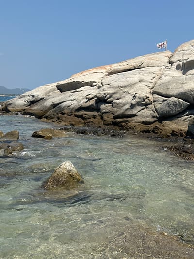 A low-angle shot captures a rocky shoreline with clear, shallow water in the foreground. The water is translucent, revealing submerged rocks and the sandy bottom. The rocks on the shore are large and weathered, with visible crevices and formations. A white flag with a red cross and black symbols is flying from a pole on top of the highest rock formation. In the background, mountains are visible under a clear blue sky. The scene appears to be a coastal area in the municipality of Castiadas, Italy. There are no people or animals visible in the image.