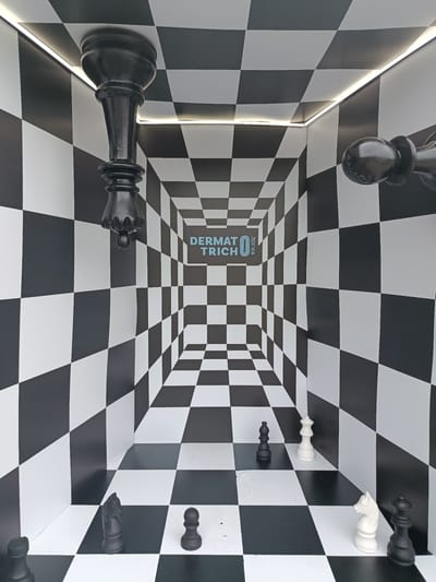 The image depicts an optical illusion room or art installation designed like a giant chessboard, located in Vynnyky, Ukraine. The scene utilizes forced perspective, with the black and white checkerboard pattern covering the floor, walls, and ceiling, converging towards a central point on the back wall, creating an exaggerated sense of depth.
Oversized chess pieces are strategically placed throughout the room. On the floor, closer to the viewer, are several black and white pieces: a black rook and a black pawn are visible on the bottom left, while a white knight and a black queen stand on the bottom right. Further into the room, a black knight and another black pawn are seen on the left side, and a white queen and a black bishop on the right. Adding to the surreal effect, large black chess pieces are affixed to the ceiling in an inverted position; a prominent black king hangs from the top-left corner, and part of another black piece, possibly a pawn or rook, is visible upside down on the top-right.
LED strip lighting runs along the seams where the ceiling meets the walls, illuminating the space and highlighting the architectural lines. On the distant back wall, centered in the converging checkerboard pattern, the light blue text "DERMATRICH" is displayed horizontally, with the word "ЛОГІКА" (Logika, meaning "logic" in Ukrainian) oriented vertically to its right. There are no people visible in the scene. The overall impression is one of an immersive, mind-bending environment playing with scale and perspective.