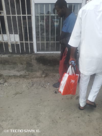 The media file shows two individuals in an outdoor setting. One person, wearing a blue shirt and red pants, is holding an orange and white shopping bag in their left hand. The other person, whose back is mostly to the camera, is wearing a white outfit. The scene appears to be in front of a building with barred windows. The ground is rough, composed of dirt and some debris.

The activity depicted is simply standing or walking, with no specific event clearly identifiable. The lighting suggests it is daytime, and the weather is not discernible from the image.

Notable details include the presence of a shopping bag with some abstract white graphic elements. A partial view of a white vehicle is visible through the barred window of the building, possibly indicating a street or roadside location. The overall impression is of a casual encounter or moment in a public space.

The visible text at the bottom of the image reads "TECNO SPARK 10c".