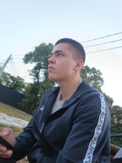 A young man with a short haircut is sitting outdoors, looking upwards and to the left. He is wearing a dark gray zippered jacket with a white stripe down the sleeve that has a repeating logo, and a light-colored shirt underneath. He is holding a black smartphone in his left hand. The background consists of trees, a power pole with wires, and a light blue sky. The image appears to be taken in a park or residential area.