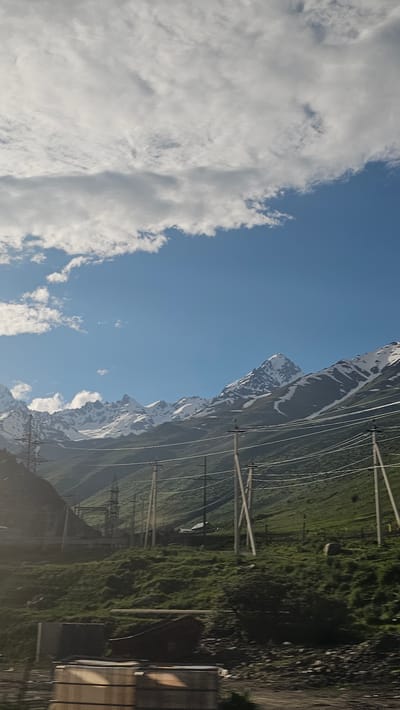 The image captures a daytime outdoor scene of a mountainous landscape, possibly near Nizhnii Zaramag, Russia, characterized by snow-capped peaks against a bright blue sky with scattered white clouds. In the foreground, a series of power lines supported by utility poles stretches across the frame, suggesting the presence of electrical infrastructure. The terrain slopes upward from the foreground, covered in green grass and dotted with some shrubs and rocks. Further back, more power lines and transmission towers are visible, indicating a continued presence of energy infrastructure. The majestic, rugged mountains dominate the background, with their snowy summits and rocky slopes. The overall impression is one of a remote, natural setting augmented by human-made structures for power transmission.