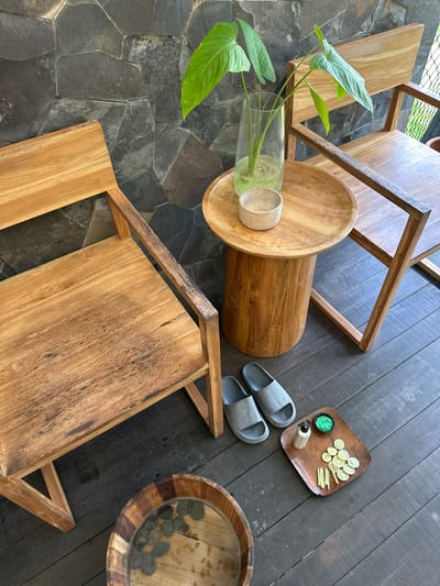 The image depicts an outdoor spa setting in Licin, Indonesia. The flooring transitions from dark stone tiles to dark wooden planks.

On the stone tiles, there is a glass vase with a green leafy plant, and a wooden coffee table.

On the wooden planks, there is a tray containing sliced lemons, lemongrass, a small jar with green contents, and a small bottle. Next to the tray is a pair of grey slippers and a wooden bowl filled with water and stones. A wooden chair is partially visible in the lower right.