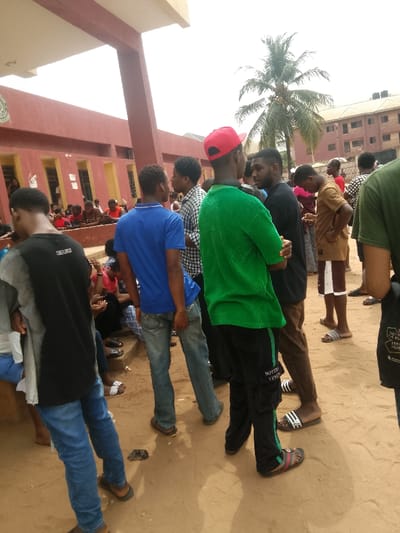 The scene depicts an outdoor gathering of approximately twenty individuals, predominantly young males, in Umuokpu, Nigeria. The ground consists of reddish-brown dirt and sand. In the left and center-left background, a large, single-story building with a reddish-brown facade and a light-colored overhead canopy is visible; several people are seated on a lower ledge or steps near this building. A tall palm tree with coconuts stands in the middle-right background, behind which a multi-story, reddish-brown building is partially visible. The sky is bright and overcast.
In the midground, facing away from the camera, a male wearing a blue polo shirt and blue jeans stands beside another male in a bright green textured short-sleeved shirt, black track pants with a green stripe and "BOTTEGA VENETA" text, and a red baseball cap. The male in green is looking towards a male in a black t-shirt and brown trousers, who has his arms crossed and is wearing black and white striped slides. To the far right, a male in a light brown short-sleeved shirt and patterned shorts is visible, looking downwards. Other individuals in the midground are standing and conversing in small groups. In the foreground, on the left, a male in a black and grey t-shirt and blue jeans is partially visible, facing away. Most individuals are wearing casual attire, including t-shirts, jeans, shorts, and open-toed footwear such as sandals and slides. The overall atmosphere suggests a casual social gathering or a waiting area.