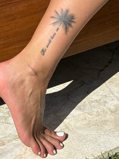 A person's left leg and foot are prominently displayed against a varied background. The leg rests on a wooden surface on the left, with a palm tree tattoo and faded script reading "This would have..." visible on the lower leg. The foot is angled towards the right, with white nail polish on the toes. A jagged shadow diagonally bisects the frame, falling across the foot and the textured, light-colored stone surface behind. Green foliage appears at the top right corner of the image. It is not possible to identify the location in the city of Medahan, Indonesia, from this image.