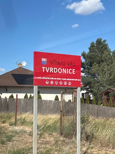 A red sign in Tvrdonice, Czechia, reads "VÍTAJÚ VÁS TVRDONICE" in white lettering, meaning "Welcome to Tvrdonice." Below this, there are several circular symbols, possibly representing local attractions or amenities. The sign is mounted on two grey posts. In the background, a house with solar panels and satellite dishes is visible, along with a wooden fence and tall green trees. The sky is blue with scattered clouds.
