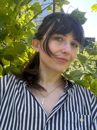 A medium close-up selfie shows a young woman with dark brown hair pulled back in a ponytail, wearing a black and white striped collared shirt. She has bangs that fall across her forehead and green eyes. A silver cross necklace is visible around her neck. She is outdoors, with lush green leaves and vines framing her face and shoulders. In the background, a light blue corrugated metal structure is visible, suggesting an outdoor setting like a garden or agricultural area. The lighting indicates it is daytime, with sunlight filtering through the leaves. The location is stated to be Zvonarevka, Russia.