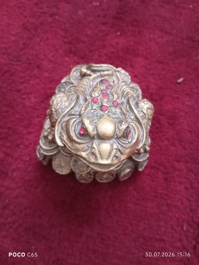 A close-up, eye-level, overhead shot shows a brass figurine of a mythical frog with red jeweled eyes and a patterned body, resting on a textured red surface. The frog's body is adorned with what appear to be small circular engravings resembling coins. The background is out of focus. The bottom left corner of the image displays "POCO C65" and the bottom right corner displays "30.07.2026 15:16". There is no discernible connection to the city of Beruniy, Uzbekistan, based on the provided image.