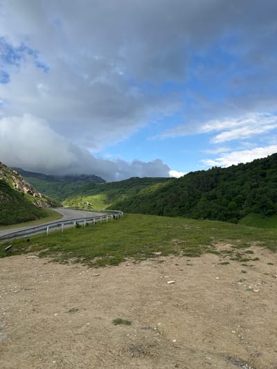The image depicts a winding road through a lush green mountainous landscape under a dramatic sky. The road curves to the left, bordered by a metal guardrail. On the left side of the road, a steep, rocky mountain slope rises, partially obscured by clouds. To the right of the road, rolling green hills covered in dense forest stretch into the distance. The foreground is a dry, dusty patch of ground with scattered stones and sparse vegetation. The sky above is a mix of blue sky and heavy, dramatic clouds, with sunlight breaking through in some areas, illuminating the scene. The location is identified as Verkhnyaya Mara, Russia. There are no people or animals visible in the image, and no explicit actions are taking place. The scene is a natural landscape with a man-made road.
