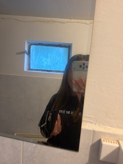 A person is taking a mirror selfie in what appears to be a bathroom. The reflection shows a white wall with a small window. The window has blue, frosted glass and is surrounded by a white frame. A crack is present on the wall near the mirror, and tiled walls are visible. The person is holding a mobile phone to take the picture, their face is obscured. They are wearing a black jacket and holding a small black purse. There is a white electrical outlet or light fixture in the top right of the mirror's reflection.

It's not possible to verify the location as Ustrem, Bulgaria based on the visual information.