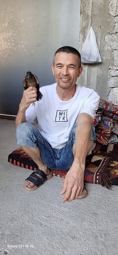 A man with short, dark hair and a light complexion sits on the ground in what appears to be a courtyard or outdoor area. He is wearing a white t-shirt with a black square logo containing "MV TR" and blue, distressed denim shorts. The man is holding a small, dark bird in his right hand, which is raised. He is smiling and looking directly at the camera.
The man is seated on a woven mat with a patterned design of red, black, and yellow geometric shapes. To his right, a pile of fabric with intricate red, blue, and green patterns is stacked against a rough, textured concrete wall. In the background, a white plastic bag hangs from a rough concrete surface. The ground is a light gray concrete. The overall impression is of an informal, outdoor setting in a warm climate. The location is identified as Ingichka, Uzbekistan.