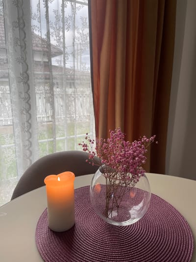A high-angle shot depicts a scene indoors, likely near a window. The composition features a white table with a round, purple woven placemat. Resting on the placemat is a lit, cylindrical candle with an orange glow. Beside the candle is a clear glass vase containing pink baby's breath flowers. In the background, a window covered with white lace curtains is visible. Part of a chair's back is also noticeable, and brown drapes are visible at the top of the frame. The location of Хлябово, Russia is not evident from the image.