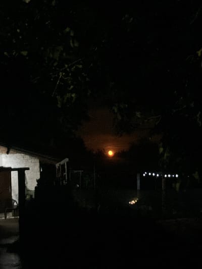 The image depicts a dimly lit outdoor scene at night, with the sky dominated by darkness and a faint orange glow in the upper center, possibly indicating a moon partially obscured by clouds or atmospheric conditions. In the foreground, a portion of a building, likely a house, is visible on the left, with a light source illuminating its exterior wall. The structure appears to be made of rough-hewn materials. To the right of the building, the landscape is obscured by shadows and foliage from overhanging trees. In the background, a line of small, bright lights, potentially decorative string lights, is visible against the dark backdrop, suggesting a garden or outdoor living area. The overall impression is one of a quiet, rural or suburban setting at night. The location is identified as Kramolin, Bulgaria. There are no visible people or animals in the image.