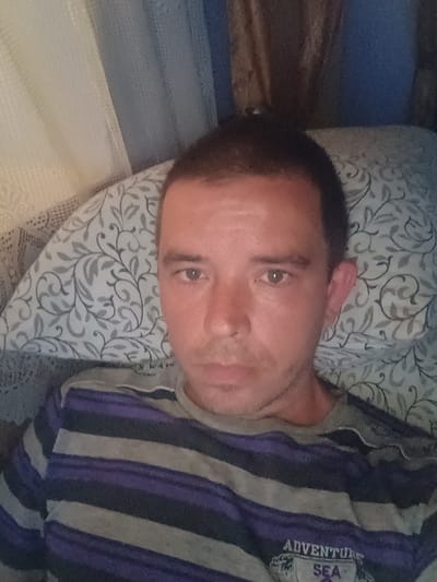 In Mitoc, Moldova, a man with short dark hair is captured in a selfie, lying on a pillow with a floral pattern. He is wearing a striped t-shirt with purple, black, and grey bands. The background suggests an indoor setting, with what appear to be curtains and a textured wall visible behind him.
