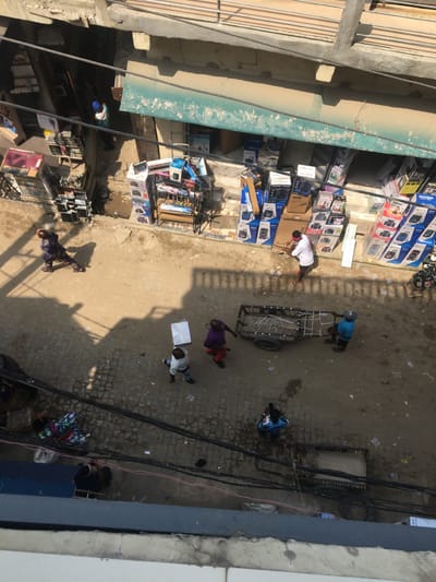 An overhead view depicts a dusty street scene in Ojo, Nigeria, under direct sunlight, casting strong shadows. The street surface is a mix of unpaved dirt and irregular cobblestone. Multiple dark electrical or communication cables traverse the upper and lower portions of the frame.
Along the right side of the street, two ground-level retail establishments operate. The building facade is light-colored and appears dusty, with a turquoise-colored awning extending over the shop fronts. Merchandise, primarily boxed electronics such as speakers and sound systems, is stacked high on display racks and directly on the ground outside the shops. A man in a white shirt and red head covering stands near the front of one shop, seemingly engaged with the merchandise. A partial view of a motorcycle is visible on the far right.
On the street, multiple individuals are present. In the midground, a man in a white shirt carries a flat, light-colored box on his head while walking from left to right. Near him, another person in a purple shirt and red pants also walks in the same direction. To their right, a third individual in a blue shirt and hat pushes a dark, two-wheeled utility cart. Further to the left, another person in a dark blue patterned outfit walks across the street.
In the foreground, a person in a blue and white patterned shirt is observed looking at a handheld device. Below this, another individual is partially visible, seated or crouched near some patterned fabric. In the top-left background, a person wearing a blue head covering and light-colored attire is visible within an entryway of another building. Litter is scattered across the street surface.