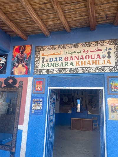 A blue wall displays a sign reading "DAR GANAOUA BAMBARA KAMLIA" in Arabic and English, surrounded by decorative swirls. Below the sign, other framed artworks are mounted, including a painting of a man playing a drum. An open doorway leads into a shop filled with various items, including musical instruments. The ceiling above has exposed wooden beams.