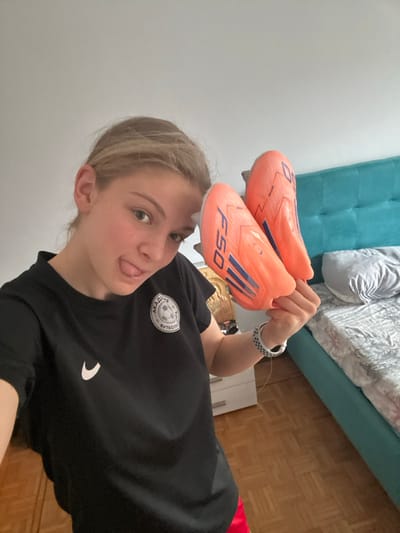 The scene takes place indoors, within what appears to be a bedroom in Roccavione, Italy. A young female, with blonde hair pulled back, occupies the center-left of the frame. She wears a black short-sleeved t-shirt featuring a white Nike swoosh on the left chest and a circular patch with Cyrillic text ('АКAДЕМIЯ ФУТБОЛУ') on the right chest. Her lower body is partially visible, showing red shorts. A white-banded smartwatch is present on her left wrist. She holds two bright orange Adidas F50 soccer cleats with her right hand, positioning them near her forehead. Her face is directed towards the viewer, with her tongue extended. The background consists of a plain light-colored wall. To the right, a bed with a teal-colored headboard and frame is visible, topped with a grey and white patterned duvet and a grey pillow. To the left of the bed, a white dresser with two silver-handled drawers stands on parquet wood flooring. A golden-colored object is partially visible atop the dresser behind the female's right arm.