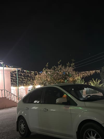 The image shows a white car parked at night, possibly in Subutli-Miatli, Russia. The car is viewed from the side, with its rear wheel visible. The scene is illuminated by strings of small, warm-toned lights that are draped across trees and a building in the background. There are also small triangular flags, some of which appear to be green, blue, and yellow, strung along with the lights. The sky is dark, indicating it is nighttime. The building in the background has arched windows, and a railing is visible in the foreground. The overall atmosphere appears festive and celebratory, possibly due to the decorations. There is no visible text in the image.