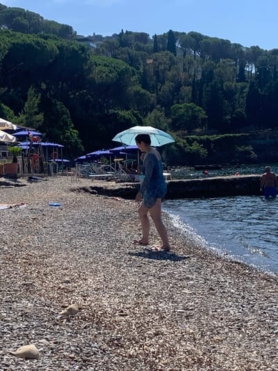 The image depicts a sunny day at a pebble beach in Pozzarello, Italy. A woman, wearing a light blue, sheer, knee-length dress and flip-flops, walks along the water's edge, holding a light blue umbrella over her head. Her dark hair is pulled back. In the background, a man stands in the water wearing blue swim trunks. The water is clear and gently laps at the shore. The beach is covered in small, light-colored pebbles. To the left of the image, there are rows of blue umbrellas and lounge chairs, suggesting a populated beach area. A lush green hillside covered in trees rises behind the beach. The sky is clear and blue.
