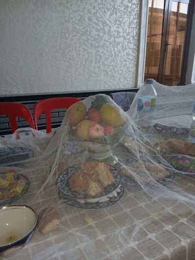 The image depicts an indoor scene, likely a dining or living area in Karshi City, Uzbekistan. A table is covered with a light-colored, sheer fabric, draped over various objects. On the table, a tiered fruit bowl is prominently displayed, filled with apples, bananas, and other fruits. Next to it, a plastic bottle of water and other items are partially visible, also covered by the fabric. In the background, two bright red chairs are visible, positioned in front of a textured light-colored wall. The overall impression is of a setting prepared for an event or meal, with food and drinks laid out and protected by the sheer covering.
