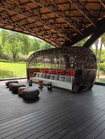 This is a horizontal, eye-level, medium shot of an outdoor seating area on a dark wooden deck, situated in Ban Pong Nuea, Thailand. The scene is bathed in bright, natural sunlight, casting subtle shadows.

The focal point is a large, curved, pod-like seating structure made of dark, woven material, resembling rattan or bamboo. This structure encloses a white, built-in sofa adorned with an assortment of decorative pillows. The pillows are a mix of solid red, and patterned designs in red, white, and black. In front of the sofa, a cluster of four rounded ottomans, each with a cushioned top, are arranged in a line on the deck. Two of these ottomans are made of the same woven material as the pod structure, while the other two appear to be crafted from wood, one lighter and one darker. Between the ottomans and the sofa, a small, wooden, stool-like table with a carved design sits on the deck, accompanied by two silver, spherical decorative objects.

The seating area is sheltered by a large, open-air roof supported by a complex, dark brown, geometric framework of beams and struts. Beyond the seating area, a lush, green rice paddy stretches out under a bright sky. The background is filled with vibrant green trees and foliage, suggesting a tropical or subtropical environment. To the right of the frame, the fronds of palm trees are visible, further indicating the location. The overall atmosphere is one of tranquil outdoor relaxation.