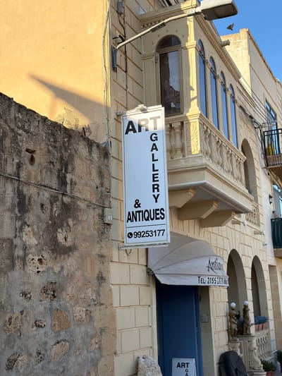 A weathered, ochre-colored building stands under a clear blue sky in Xagħra, Malta. On the left, a rough, stone wall with gaps and textures shows signs of age. A shadow stretches diagonally across the wall from an unseen object. A white sign with black lettering reads "ART GALLERY & ANTIQUES" and displays the phone number "99253177". The building itself is adorned with a decorative balcony featuring ornate railings and columns. Below the balcony, a white awning with the word "Antiques" visible is stretched over a dark blue door. Another sign, partially visible at the bottom, also reads "ART G A". The scene is illuminated by bright sunlight, casting sharp shadows.