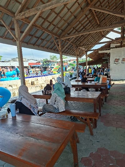 The image shows a scene of people sitting at wooden tables and benches under a wooden roof structure. The tables and benches are arranged in rows, extending into the distance. Several people are seated at the tables. Some people are wearing headscarves. In the distance, there are a group of people gathered around a pool of water. The scene appears to be a public space, perhaps an outdoor dining area or a park. The location is in Kecamatan Tulung, Indonesia, with coordinates -7.592 latitude and 110.65 longitude.