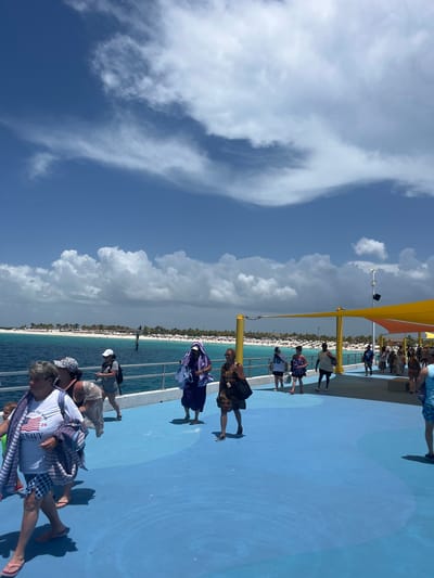 The image shows a bright, sunny day on a pier in the City of Freeport, Bahamas. A group of people are walking along a bright blue, wavy-patterned surface. In the background, the turquoise ocean meets a white sandy beach lined with palm trees and resort buildings. Several small boats and umbrellas are visible on the beach. The sky is a vibrant blue with scattered white, fluffy clouds. The people on the pier are dressed in casual, summery clothing, with some carrying bags or towels. The pier itself has a metal railing along the side, and part of the pier is covered by a bright yellow awning. The overall scene depicts a lively tropical vacation destination.
