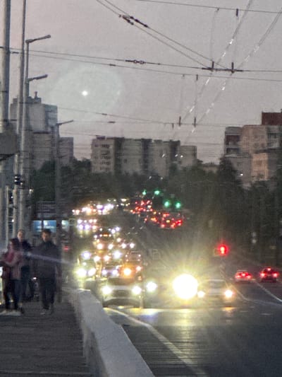 The image depicts a street scene at dusk or dawn in Rodnichkovoye, Russia. The foreground features a pedestrian walkway with a white railing. Several individuals are walking on the walkway, appearing to be dressed casually. Beyond the walkway is a road filled with traffic, characterized by numerous car headlights and taillights. The vehicles are illuminated by their own lights, creating a bright and somewhat blurry effect due to the low light conditions and potential motion blur. Traffic lights are visible, with one appearing red. In the background, there are buildings and trees, with the sky overcast and a bright, hazy light source, possibly a street lamp or the sun peeking through clouds, creating a lens flare effect. Overhead, there are power lines and infrastructure associated with urban transportation, such as trolley wires. The overall impression is of a busy urban environment transitioning from day to night.