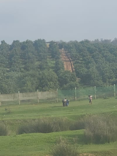 The image displays an outdoor, rural-like scene in Ameur عامر, Morocco, under an overcast or hazy sky. The foreground consists of uneven terrain covered with green grass and scattered clumps of taller, dry, brownish-green brush.
In the midground, a flat, open expanse of green grass dominates, bounded horizontally by a low concrete fence with several vertical support posts. Behind this concrete structure, a section of green mesh fencing is partially visible. Within this grassy area, two distinct groups of individuals are present. To the left, a cluster of approximately three to four individuals, appearing to be children or adolescents, are gathered closely. One individual, dressed in dark blue, is positioned low to the ground, possibly crouching or interacting with an object. The other individuals are standing upright in close proximity. Further to the right, a single individual, appearing to be a child, stands with both arms raised, wearing patterned clothing.
The background is dominated by a densely vegetated hillside or forested slope, composed of numerous green trees. A light brown dirt path or track ascends this slope, creating a visible, less vegetated strip through the trees. The sky above is a uniform, light grey.