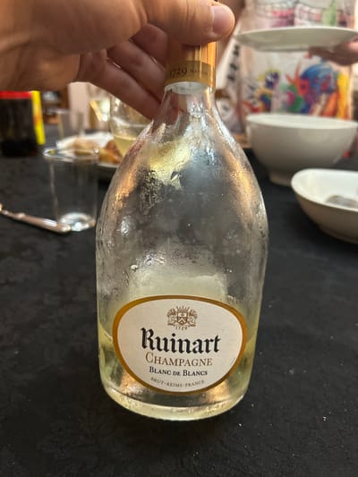 A light-skinned hand grips the neck of a nearly empty, clear glass bottle of Ruinart Champagne Blanc de Blancs. The bottle, which contains a small amount of pale yellow liquid at its base, is covered in condensation, indicating it was recently chilled. Its white and gold label prominently displays "Ruinart CHAMPAGNE Blanc de Blancs BRUT - REIMS - FRANCE" along with a crest. The bottle's gold foil neck wrap shows "1729 MAISON." The scene is set indoors on a dark, patterned tablecloth. To the left, an empty clear glass tumbler and a silver fork are visible. The blurred background shows a social gathering with several white ceramic bowls, another glass containing a pale liquid, and a person in the upper right holding a white plate, wearing a garment with colorful, abstract patterns. This event is located in the city of Mogadouro, Portugal.