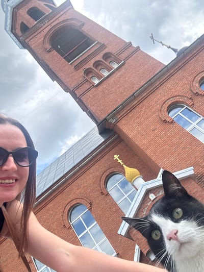A low-angle, selfie-style photograph captures a woman and a black and white cat in front of a red brick church in Medvedevo, Russia. The woman, wearing sunglasses and a black top, is on the left side of the frame, her arm reaching out. Her mouth is open in a smile. To the right of the woman, a black and white cat with green eyes is prominently featured, looking directly at the camera. The cat's ears are perked up. Behind them, the tall, red brick bell tower of the church dominates the background. Architectural details of the church, including arched windows and a golden dome with a cross, are visible. The sky is cloudy.