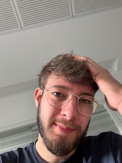 A close-up, upward-angled shot depicts a male subject from the chest up, positioned centrally in an indoor environment. The subject has short, wavy brown hair, a full beard, and is wearing gold-framed, hexagonal-shaped eyeglasses. His eyes are brown, and he displays a slight, closed-mouth smile. His right hand is raised, with fingers partially obscured by his hair, touching the right side of his head. He is wearing a dark blue or black t-shirt. The background consists of a plain light grey wall on the left and a white wall on the right, which appears to be part of a window frame or door frame, allowing diffused light from the right. The top portion of the image shows a suspended ceiling with square white acoustic or ventilation panels arranged in a grid pattern. There is no discernible information within the image to specify the exact location within the city of Lièpvre, France.