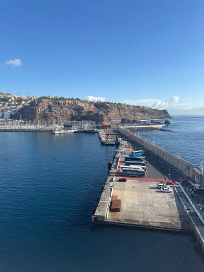 The image depicts an elevated view of the port of San Sebastián De La Gomera, Spain, under clear, bright daylight. In the foreground, a large, rectangular concrete pier extends into the deep blue water. On this pier, several blue passenger buses are parked in a row, accompanied by a red car and multiple white vans or cars further down. Rust-colored rectangular objects are visible on the concrete surface. Numerous individuals are scattered across the pier and a connecting walkway, suggesting pedestrian activity.
To the left of the main pier, a marina is densely packed with white sailboats and other recreational vessels. A small yellow boat or ferry is docked near the entrance to this marina. A long, grey concrete breakwater or jetty, equipped with light poles, extends into the sea, forming the outer boundary of the port area, with a visible road running along its length.
The background features the town of San Sebastián De La Gomera, built upon steep, rugged, reddish-brown cliffs and hillsides. Residential buildings are densely clustered across these natural formations. The sky above is a clear, bright blue with a few wispy white clouds. The horizon in the far distance reveals a faint landmass, possibly another island. The water within the harbor is calm and deep blue.