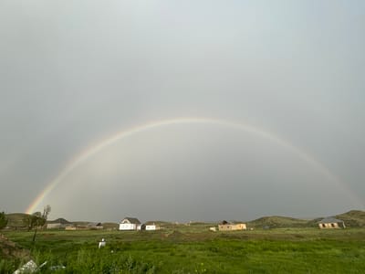 The image shows a wide, panoramic view of a rural landscape with a prominent rainbow arching across a gray, overcast sky. In the foreground, there is a vibrant green grassy field, with some patches of yellow wildflowers. Several houses are scattered throughout the middle ground, varying in size and architectural style, some appearing to be in various states of completion. The houses are situated amidst rolling hills that form the backdrop of the scene. The sky is predominantly gray, indicating recent or ongoing rain, which has likely caused the rainbow. The overall impression is of a tranquil, remote setting in the village of Kara-Oi, Kyrgyzstan, after a rainfall.