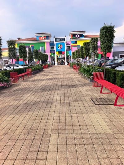 A bright, colorful outdoor shopping village is depicted. The buildings are painted in vibrant, geometric patterns of pink, green, yellow, blue, and orange, with a distinctive multicolored circular mosaic above the central entrance. The sky is partly cloudy, suggesting it could be midday. A paved walkway, made of light brown square tiles, leads towards the shops, lined with red benches and lush green trees and flowering plants. Several cars are parked in the background, indicating this is a commercial area. A sign above the entrance reads "Palmanova designer village". The overall atmosphere appears cheerful and welcoming, with a few people visible walking around, hinting at a typical day at a retail outlet.
